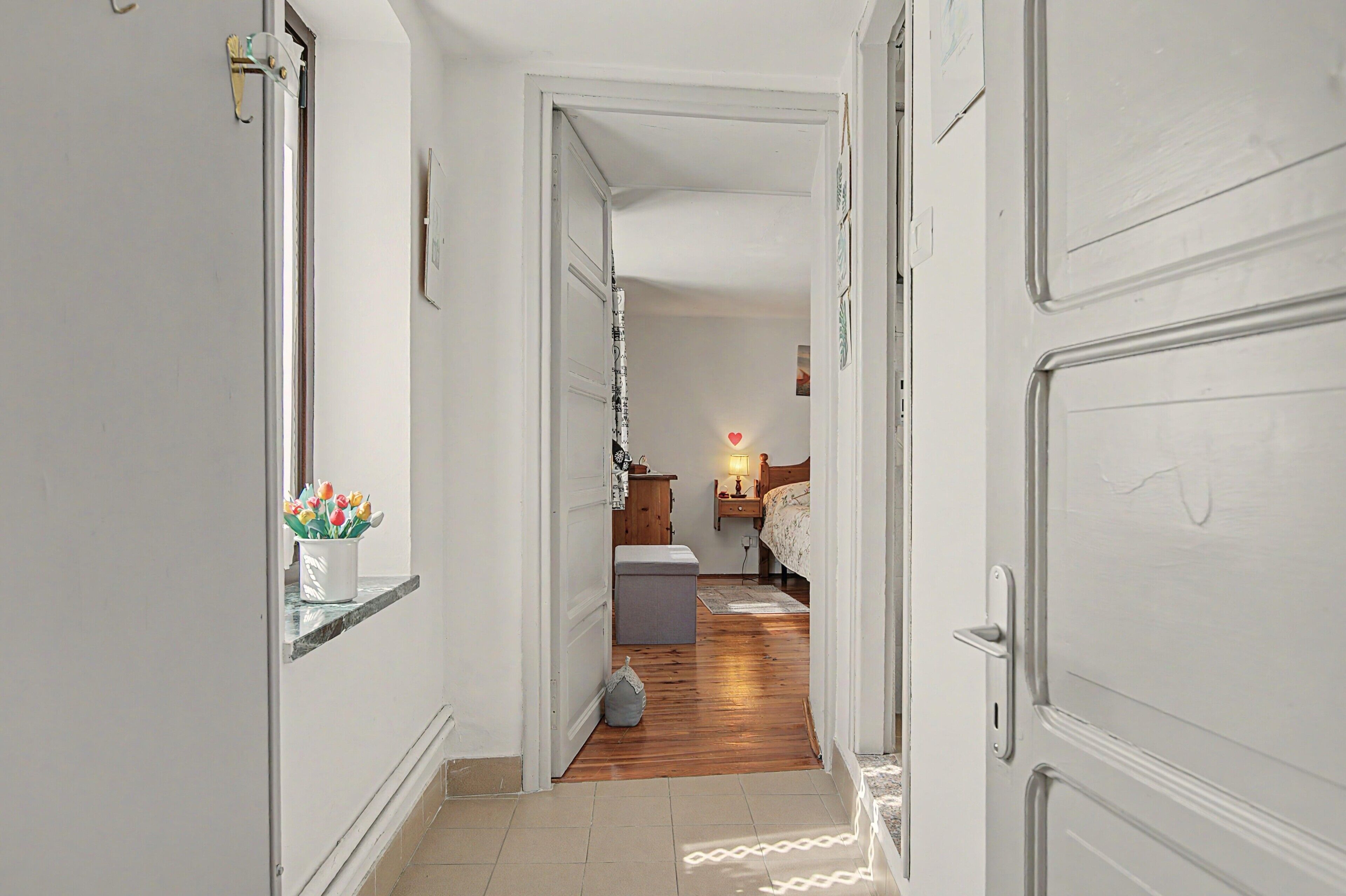 Un couloir mène à une chambre à coucher qui contient un lit, une table de chevet, une lampe et une petite commode.