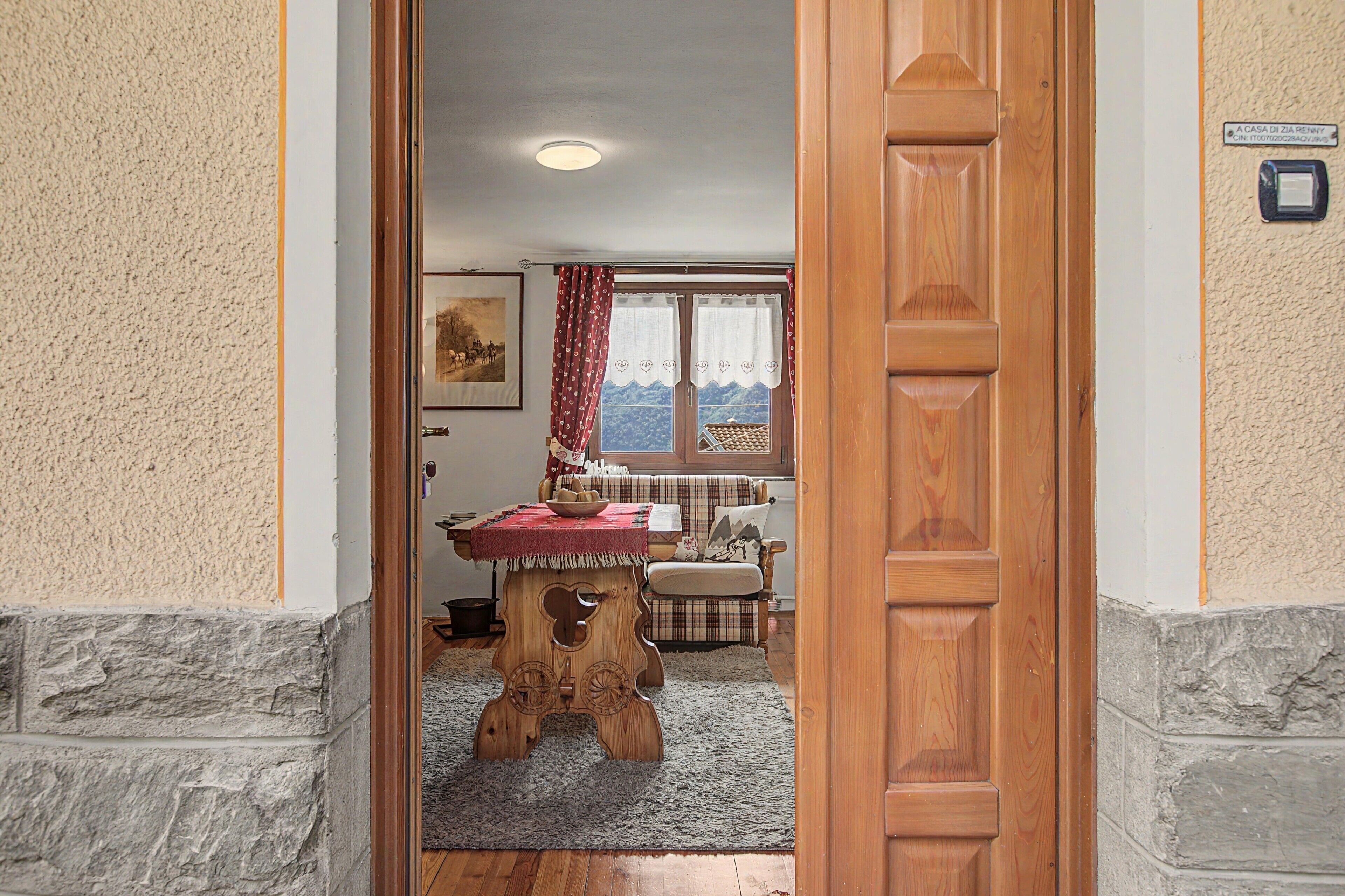 Puerta de madera que da a un cuarto con una mesa de madera, un banco y vistas al exterior a través de una ventana con cortinas.
