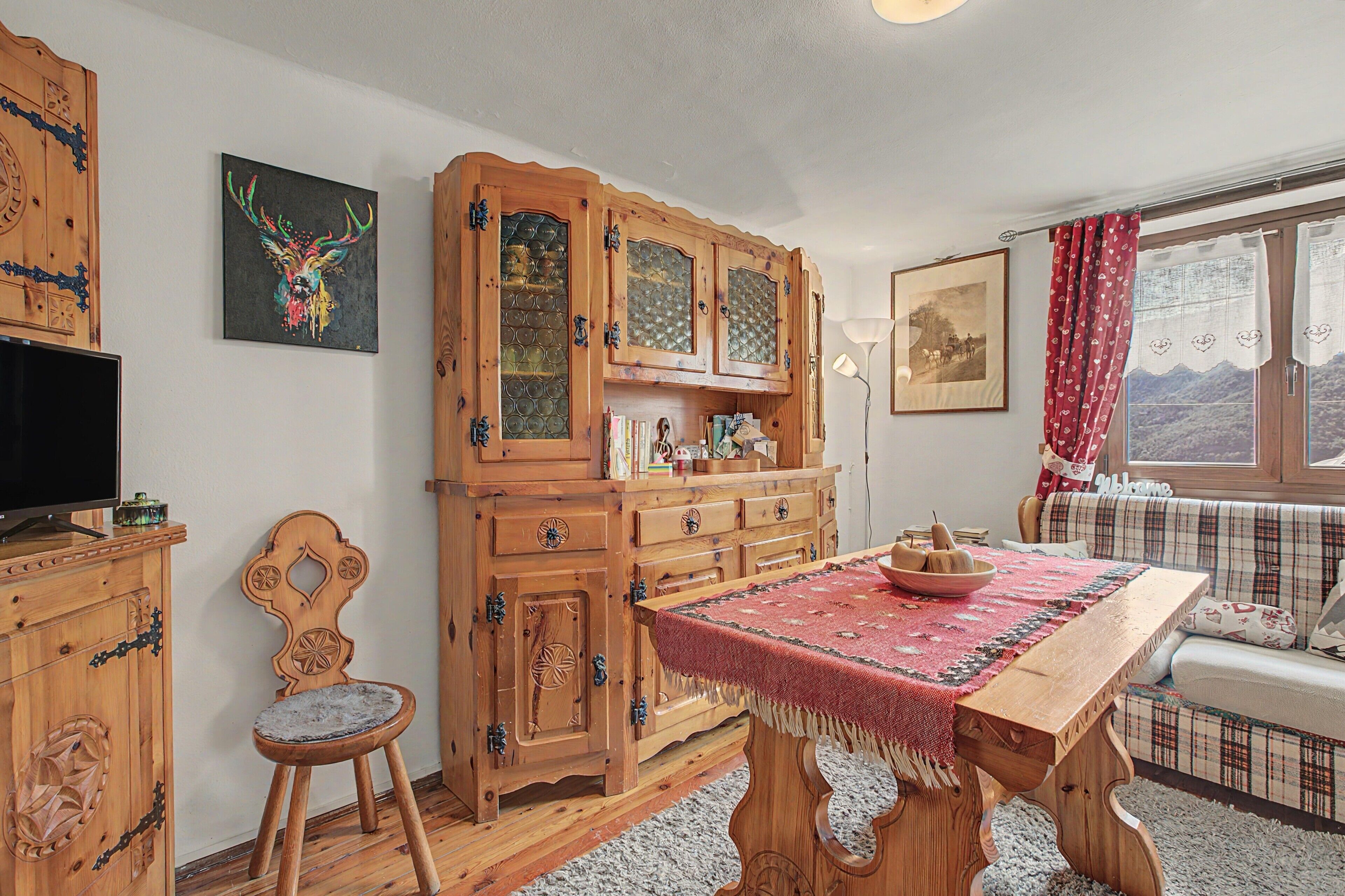 Una habitación acogedora con una mesa de comedor de madera, una silla, un aparador con puertas de cristal, un televisor y una ventana con cortinas.