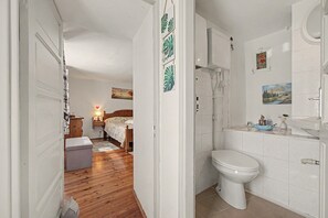Une chambre avec un plancher en bois, un lit recouvert d’une couvre-lit à motifs floraux, une table de chevet avec une lampe et un mur orné d’un tableau encadré.