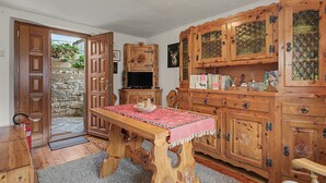 Une table à manger en bois recouverte d’une nappe rouge, entourée de meubles en bois dotés de portes en verre, d’un téléviseur et d’un mur en pierre à l’extérieur.