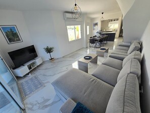 A modern living room with a flat-screen TV, a dining area, and a kitchen in the background. - Villa Catherine, Paphos 150 metres from Venus Beach – come and visit us. (PAFOS)