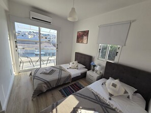 A bedroom with two beds, a window, a ceiling fan, and a view of the cityscape. - Villa Catherine, Paphos 150 metres from Venus Beach – come and visit us. (PAFOS)