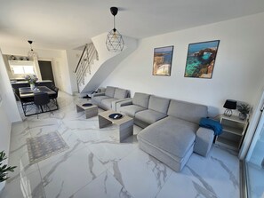 A modern living room with a grey sectional sofa, a dining table with chairs, a staircase, and two framed pictures on the wall. - Villa Catherine, Paphos 150 metres from Venus Beach – come and visit us. (PAFOS)