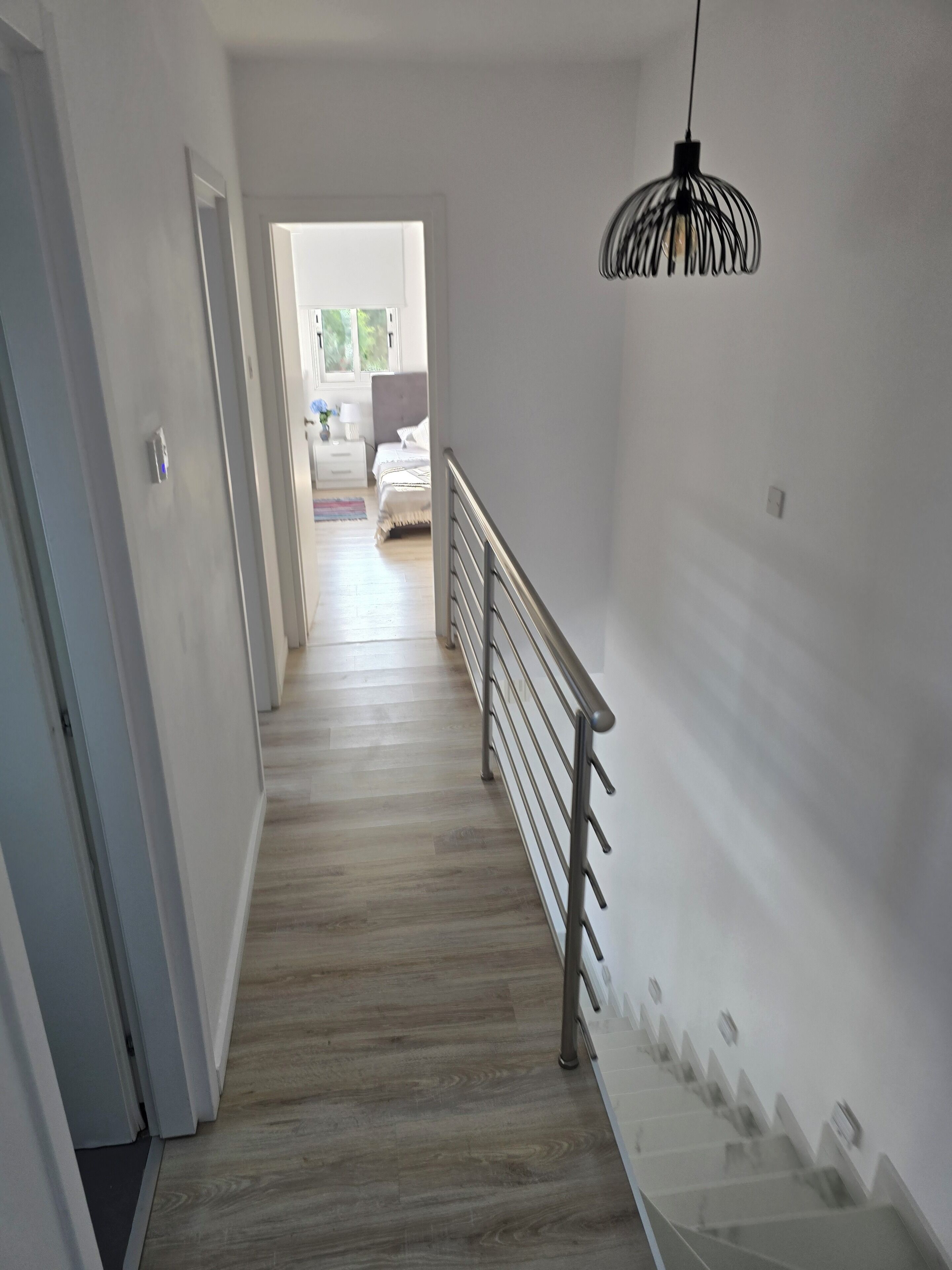 Un couloir doté d’un plancher en bois, d’une porte blanche, d’une rambarde en métal et d’un luminaire suspendu.