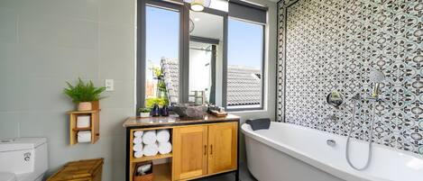 Une salle de bain moderne avec une baignoire sur pieds, un meuble-lavabo en bois, une grande fenêtre et un mur de douche orné d’un motif.