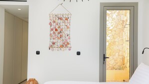 A bedroom with a bed, a wall-mounted macrame piece, a door leading to a stone wall, and a small wicker basket. - Villa Noema, Luxury Villa (Corfu)