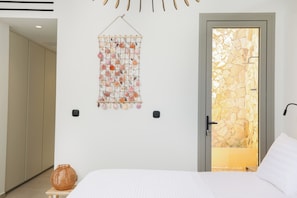 Ein Schlafzimmer mit einem Bett, einer Wanddekoration aus Makramee, einer Tür zu einer Steinwand und einem kleinen Korbwäscheschale.