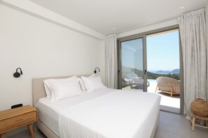Ein Schlafzimmer mit einem großen Bett, einem Nachttisch, einem Sessel und Blick auf einen Balkon mit Gartenmöbeln.