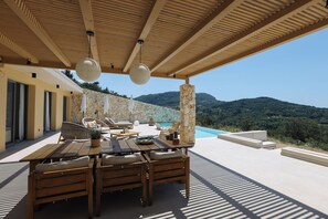An outdoor patio with a wooden table and chairs, a stone wall, and a view of a mountainous landscape. - Villa Noema, Luxury Villa (Corfu)