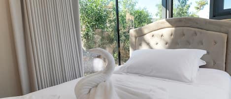 A neatly made bed with a swan-shaped towel and a pillow, a window with a view of greenery, and a curtain.
