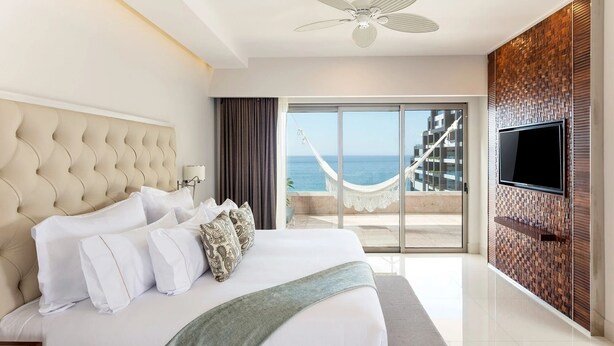 Ein Hotelzimmer mit einem großen Bett, einem Fernseher, einem Balkon mit Meerblick und einem Deckenventilator.