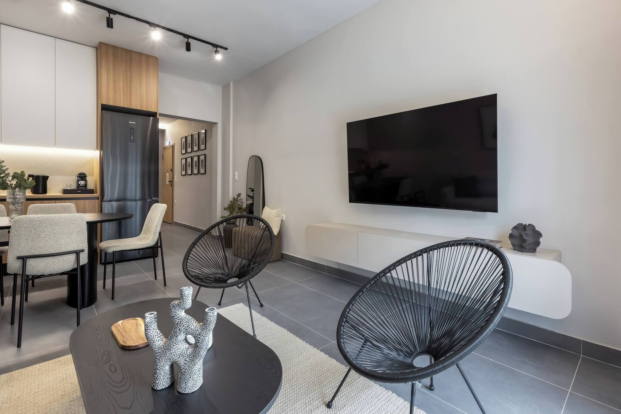 A modern living room with a dining area, a large flat-screen TV mounted on the wall, and a kitchen in the background.