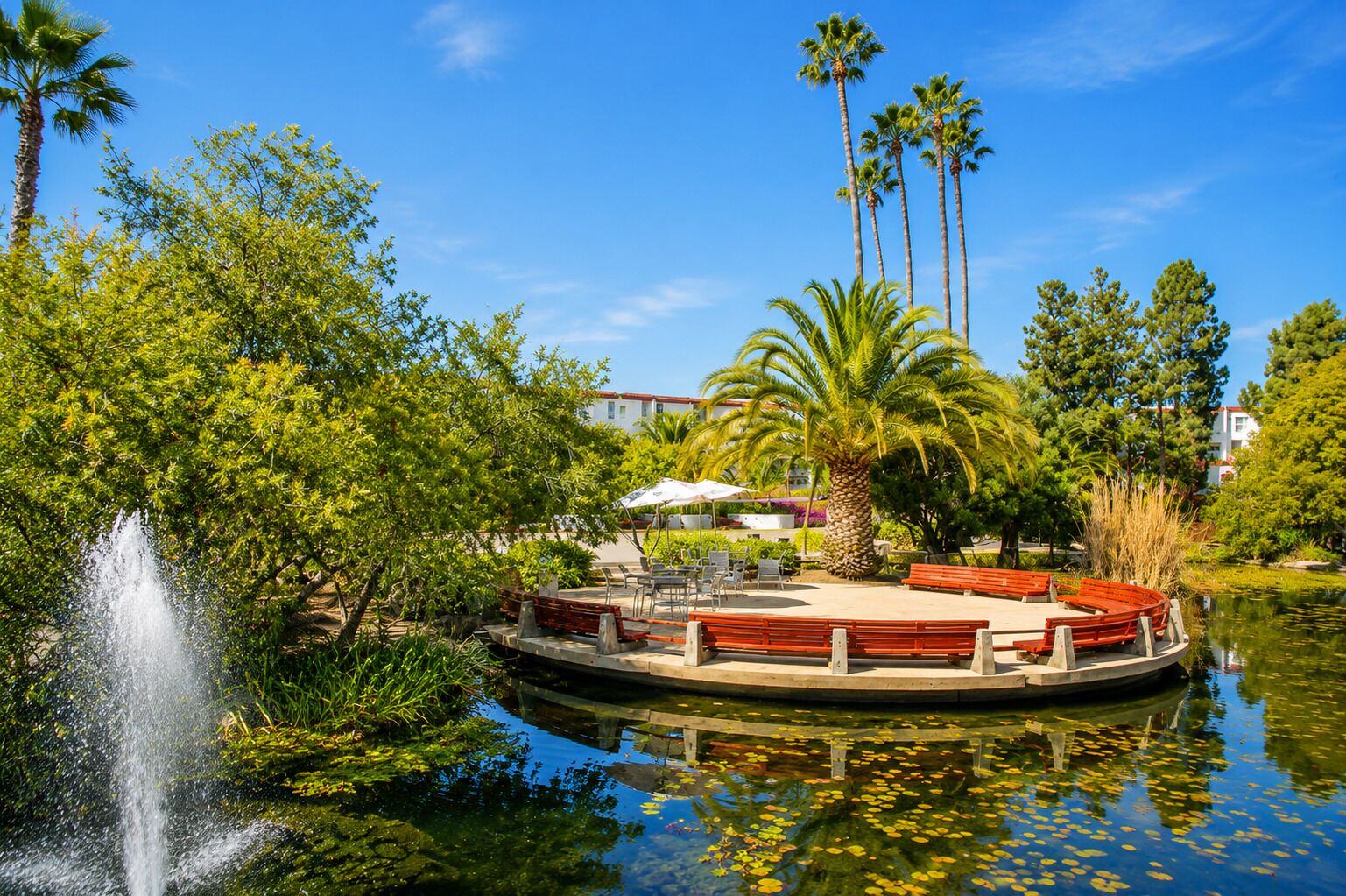 A serene water body with a fountain, surrounded by lush greenery and a wooden boat dock.