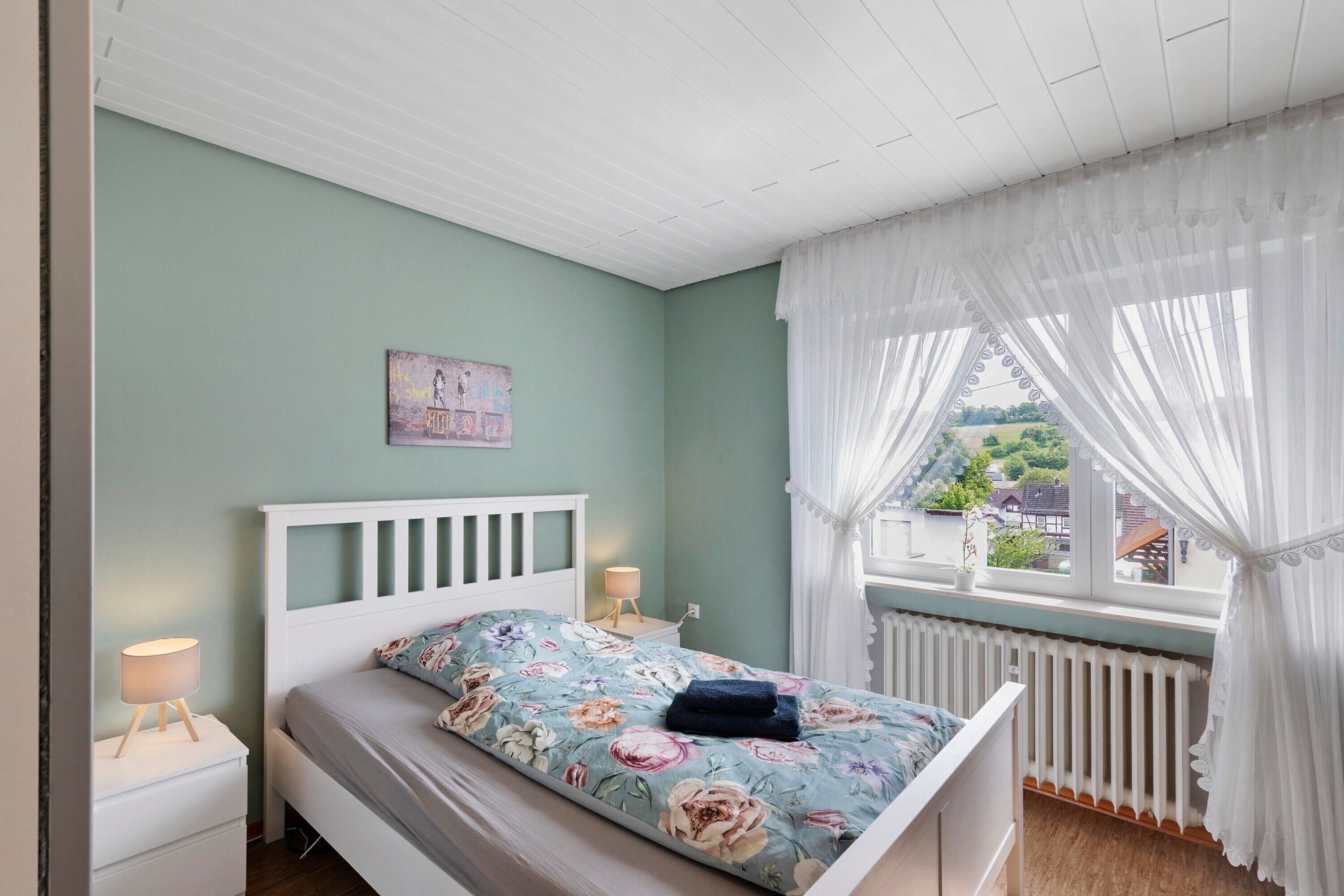Un dormitorio con cama, mesita de noche, ventana con cortinas y vistas al exterior.