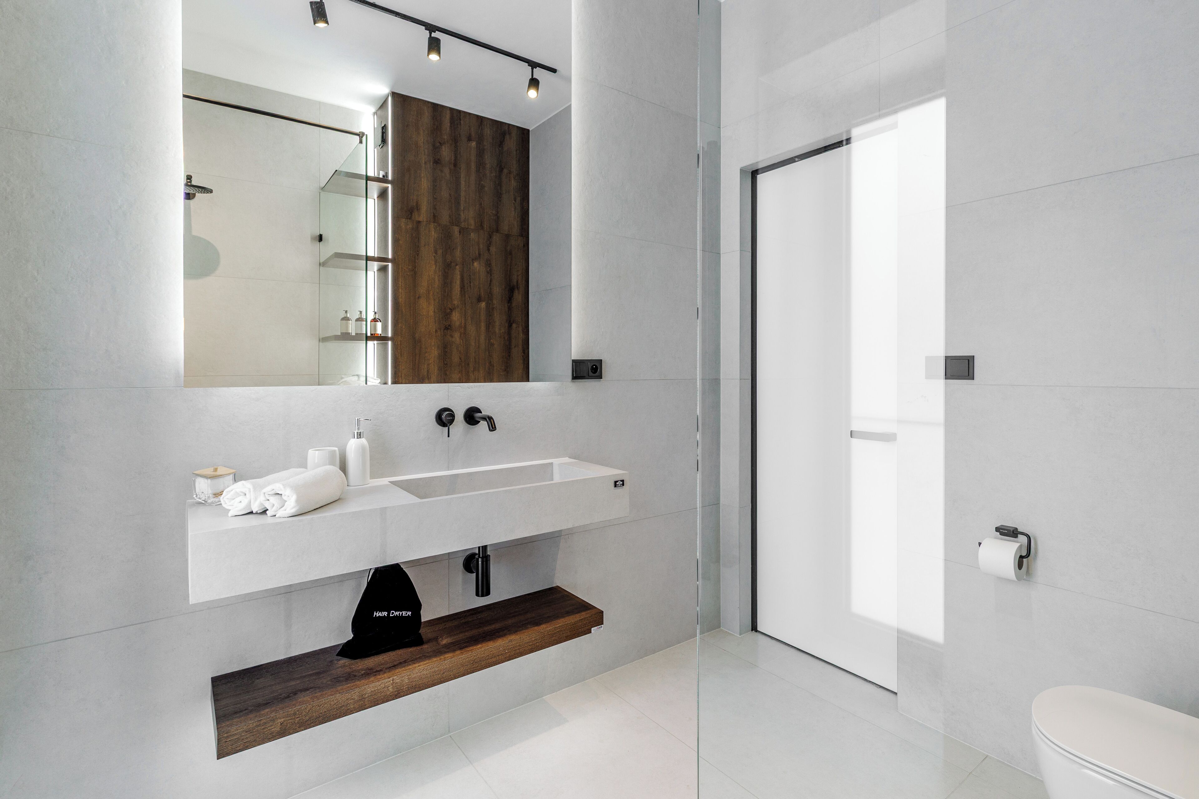 A modern bathroom with a white sink, mirror, and a wooden shelf.