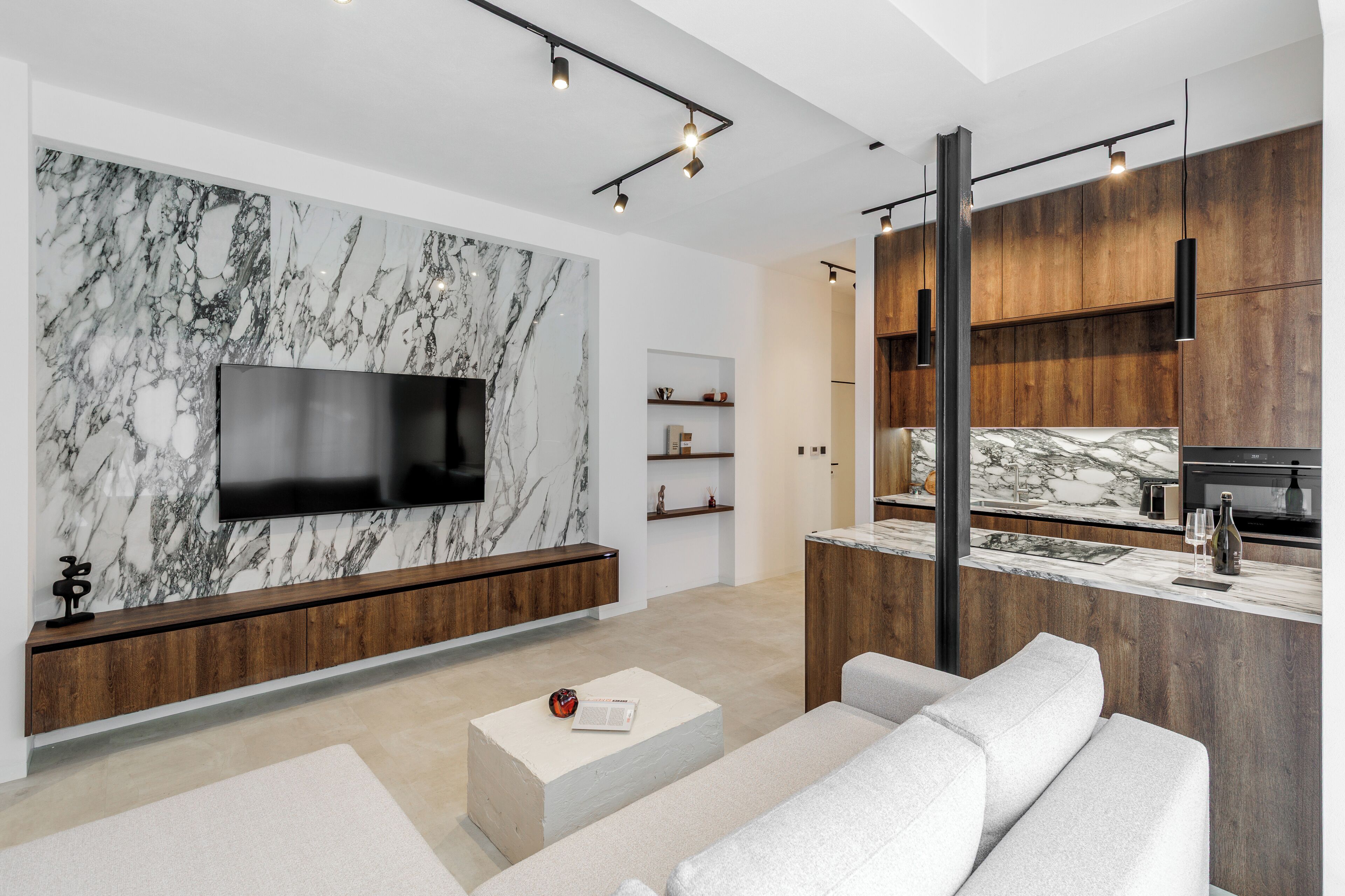 Una sala moderna con pared de mármol, televisor de pantalla plana, consola de madera, sofá blanco y una zona de cocina con armarios de madera y encimera de mármol.