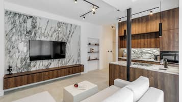 Una sala moderna con pared de mármol, televisor de pantalla plana, consola de madera, sofá blanco y una zona de cocina con armarios de madera y encimera de mármol.