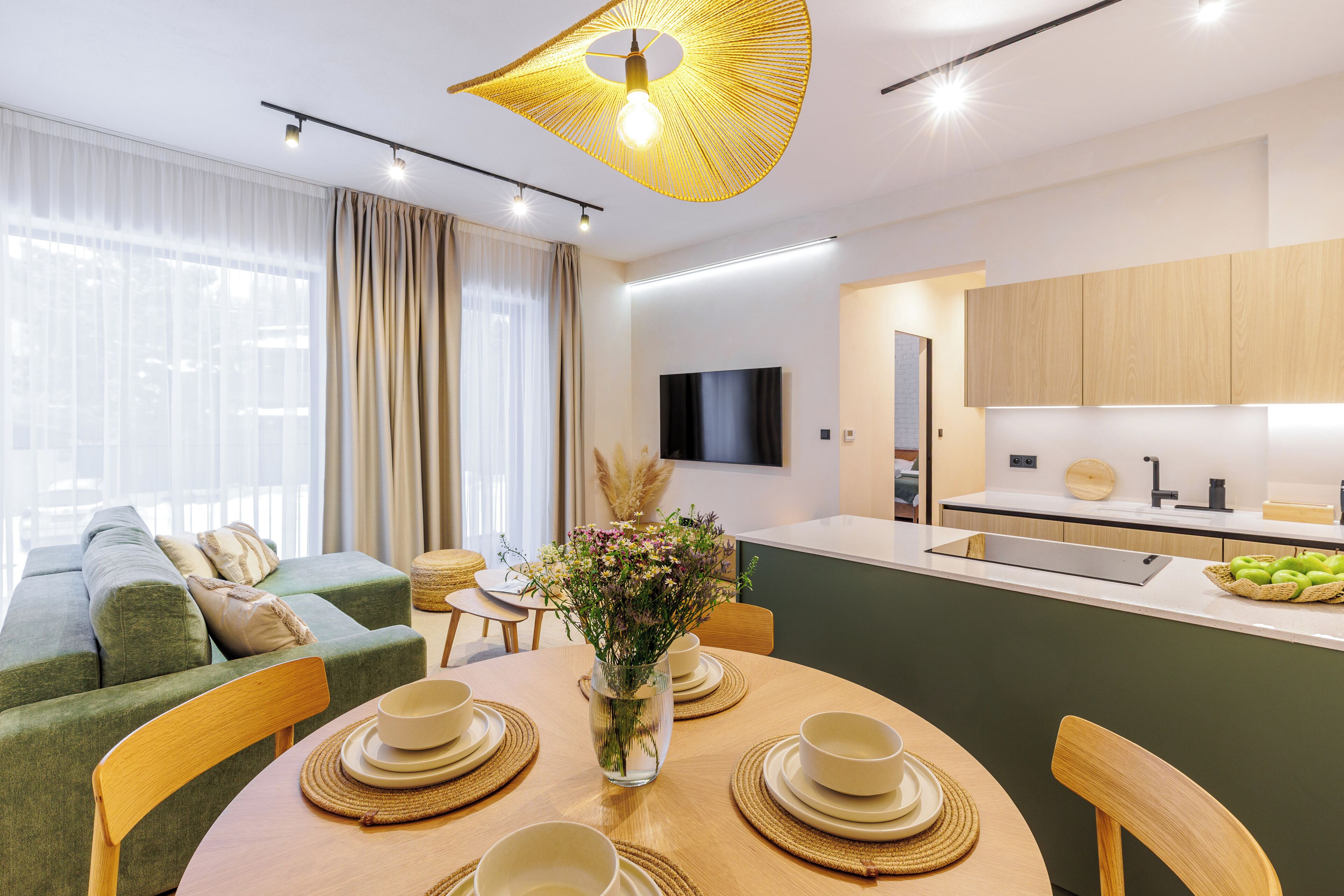 A modern living room with a sofa, a dining table set with bowls and a vase of flowers, and a kitchen area with a flat-screen TV and wooden cabinets.