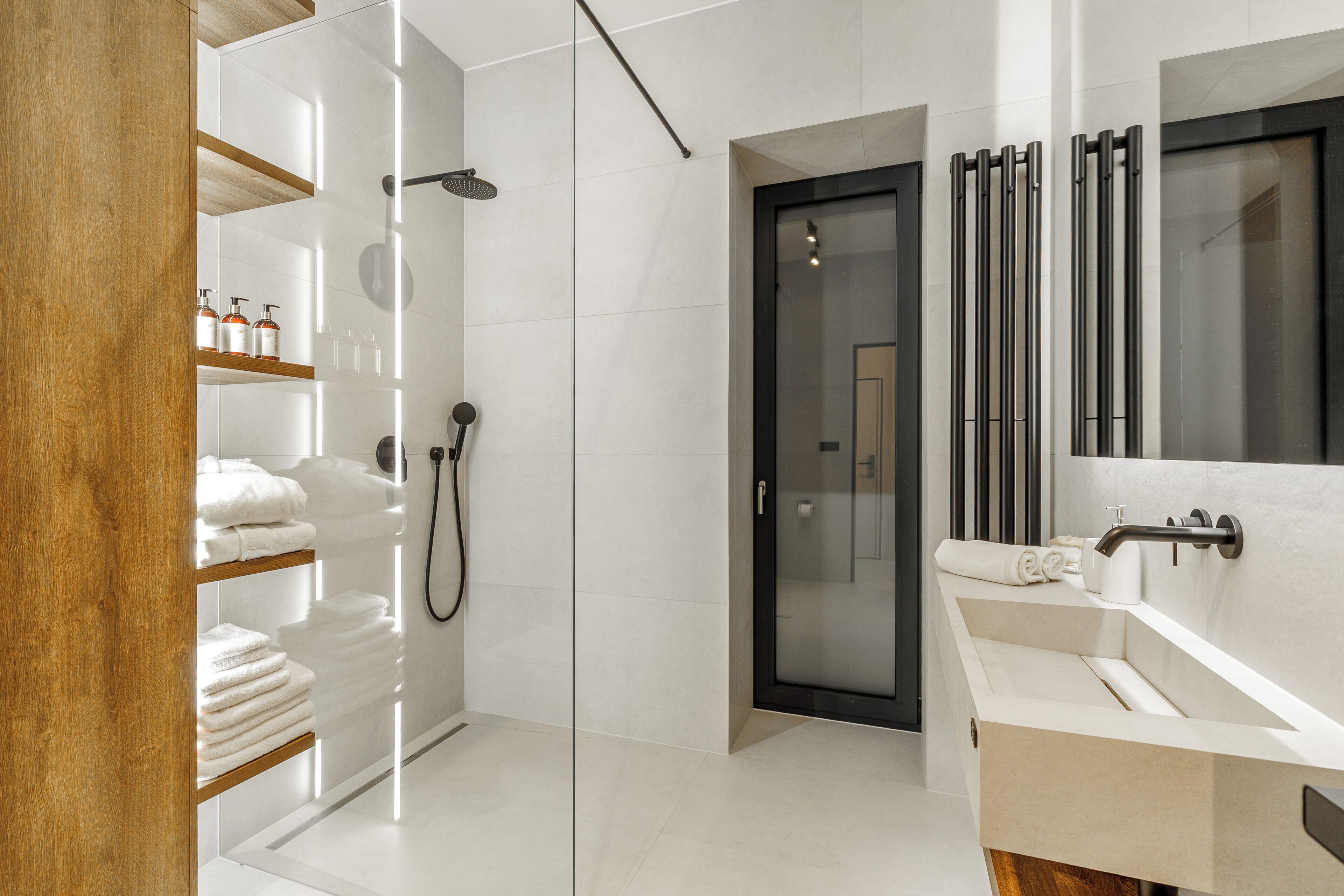 A modern bathroom with a glass shower enclosure, a wooden shelf with towels, a white sink, and a large mirror.