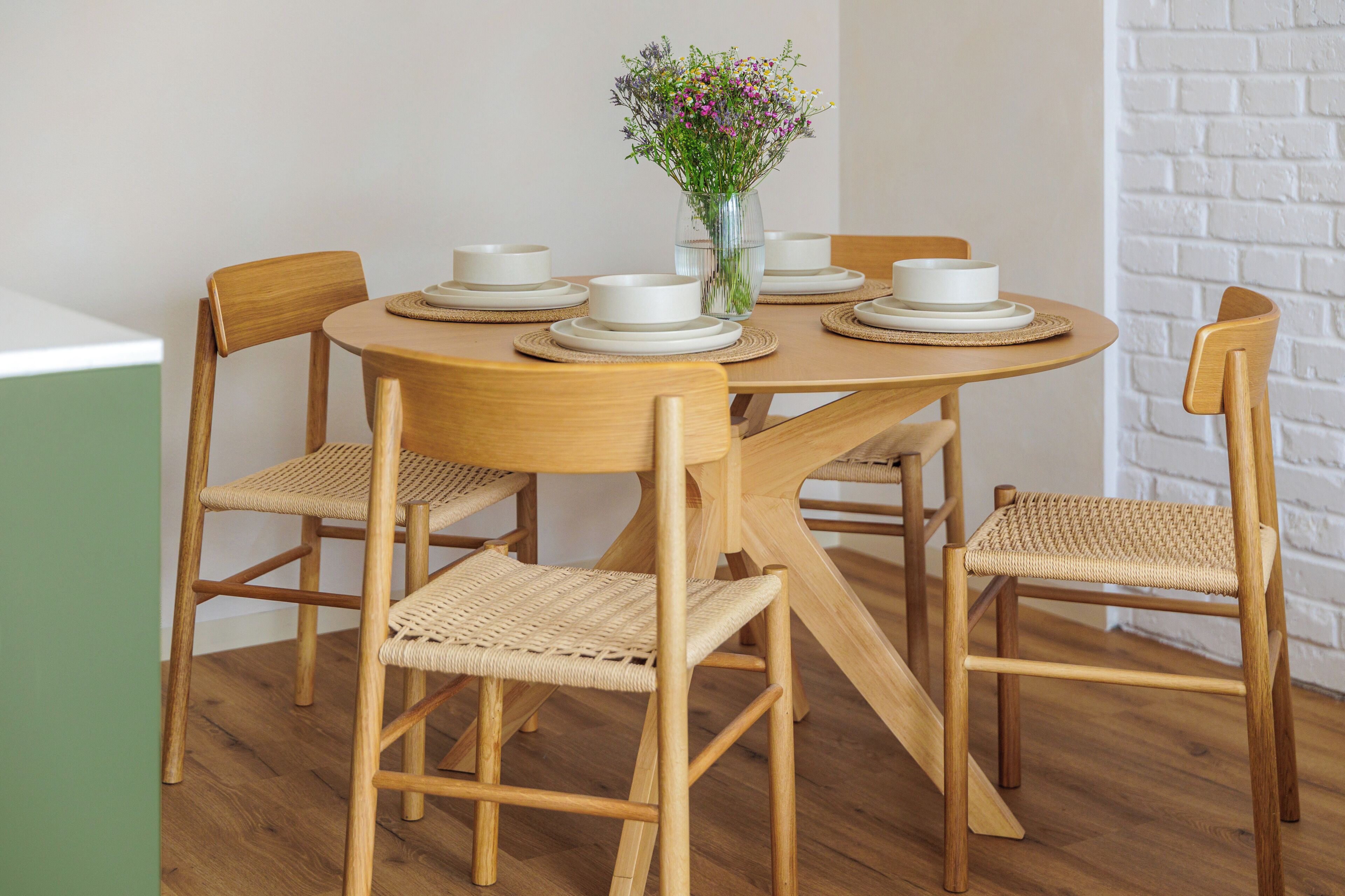 A round wooden dining table with four chairs, each with woven seats. A vase with flowers is placed in the center of the table.