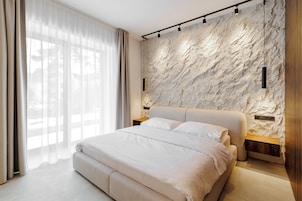 Quarto moderno com parede estampada em mármore, uma cama grande com roupa de cama branca e uma janela com cortinas translúcidas.