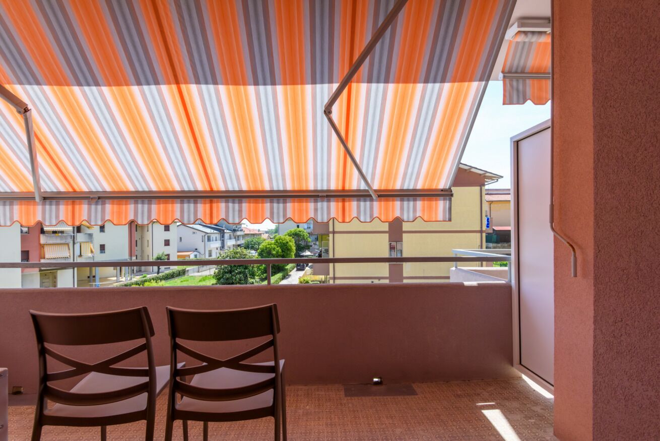 Un balcón con toldo a rayas, dos sillas y vistas a edificios.