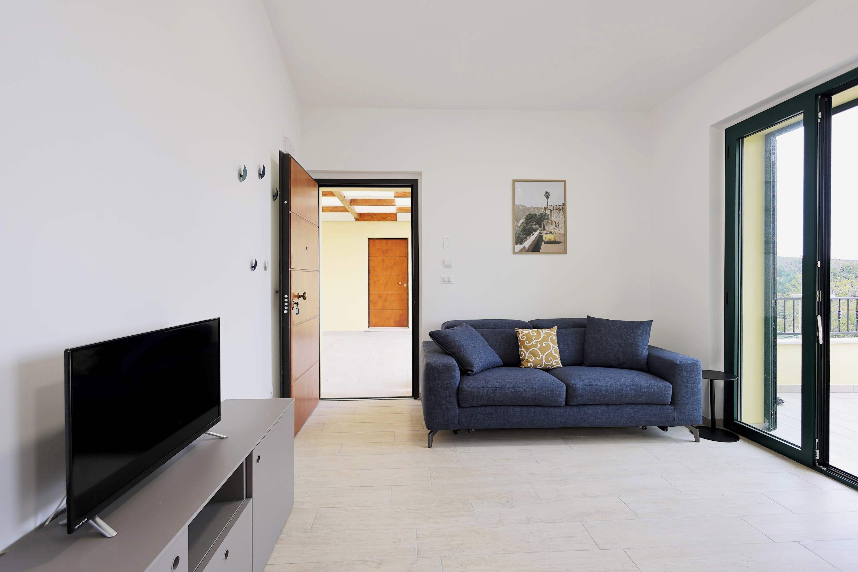 A modern living room with a flat-screen TV, a sofa, and a sliding glass door leading to a balcony.