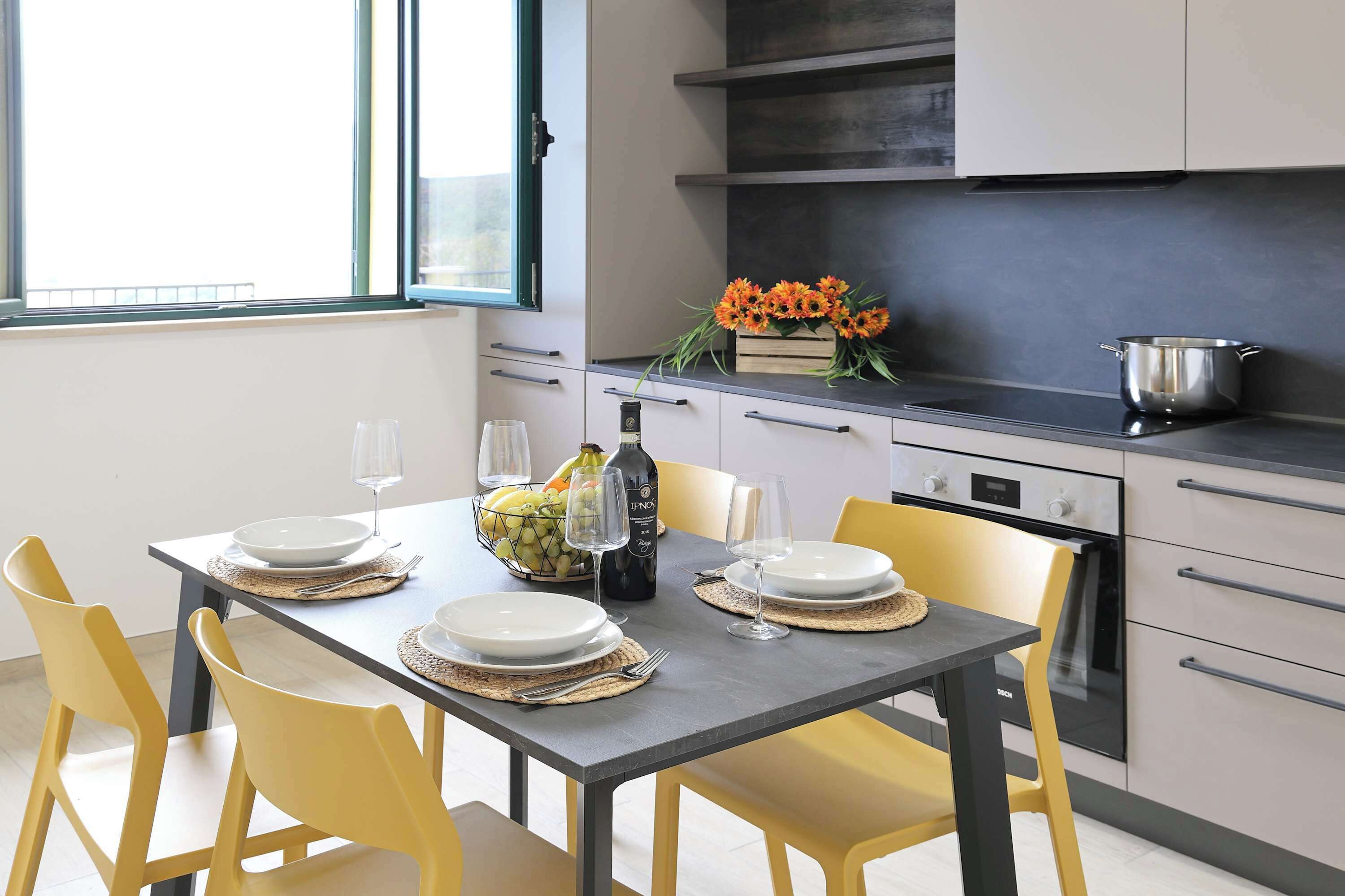 A modern kitchen with a dining table set for two, a fruit bowl, wine bottle, and a flower arrangement.