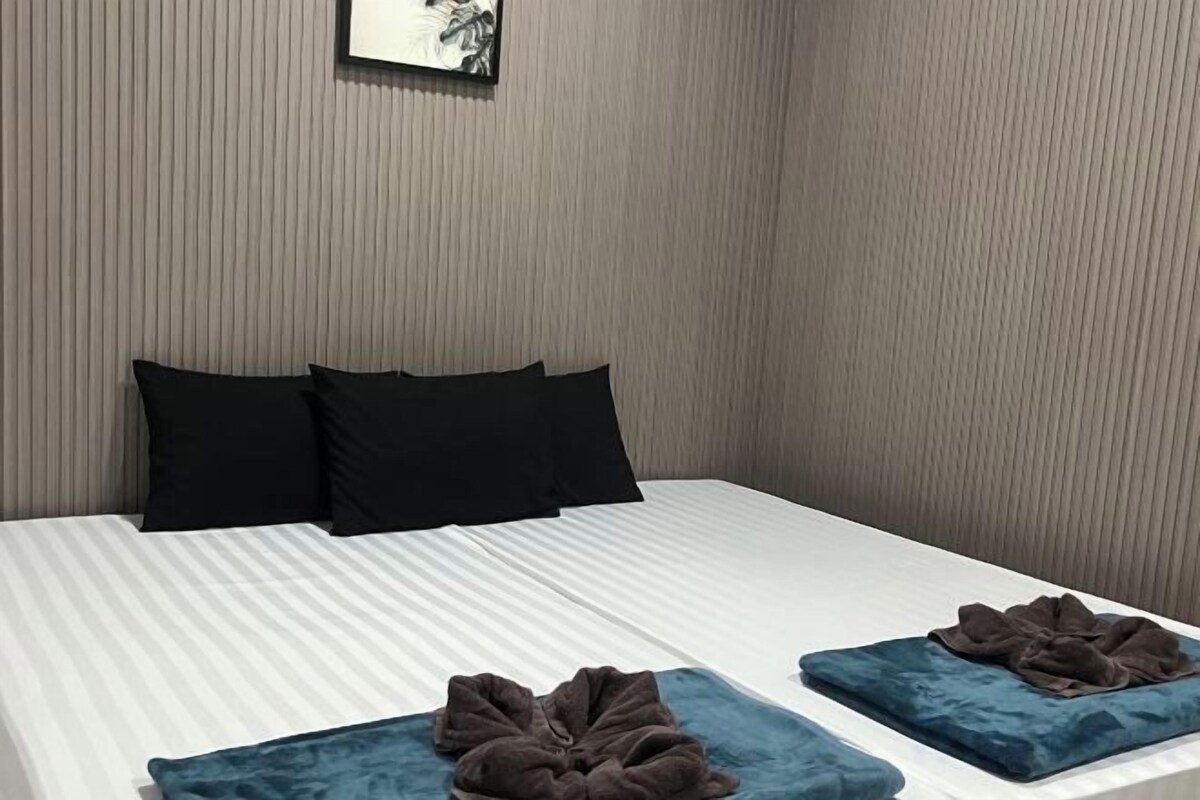 A double bed with a white sheet and two dark pillows, a blue blanket, and a blue throw pillow on a white nightstand.