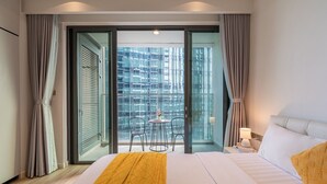 Habitación de hotel con cama, una pequeña mesa con sillas, un balcón con vistas a un edificio y una mesita de noche con lámpara.