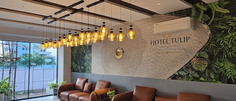 A modern hotel lobby with a reception desk, leather sofas, hanging pendant lights, and a mural with the hotel name.