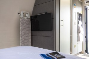 A hotel room with a bed, a television mounted on the wall, a towel rack, and a wardrobe. - Luxury Pod With Wave Pool Views & Smart Features (Newbridge)