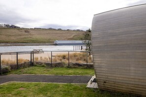A waterfront view with a large cylindrical building, a picnic table, and a fence. - Luxury Pod With Wave Pool Views & Smart Features (Newbridge)