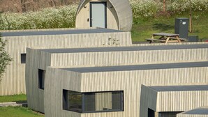 A modern building with a unique curved roof, a small enclosed area with a door, and a grassy area with people and kayaks.