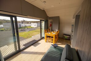 A modern room with a large sliding glass door, a dining table with chairs, a sofa, and a small kitchenette. - Waterfront Wave Cabin at Lost Shore Surf Resort (Newbridge)