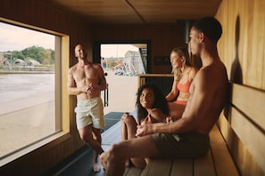 A sauna room with wooden walls, a window, and a view of a beach and buildings outside.