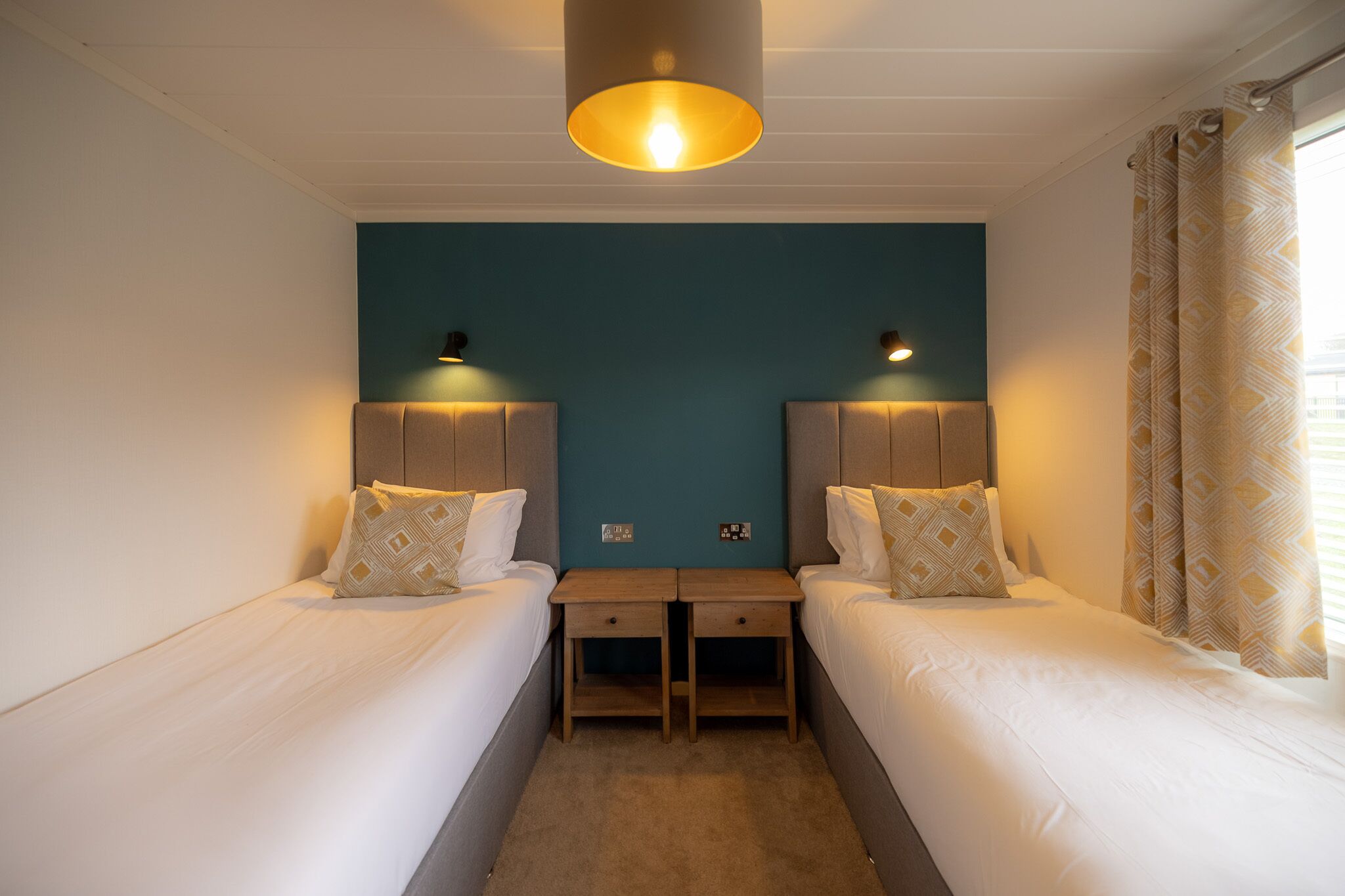 A hotel room with two single beds, a small desk, a ceiling light, and a window with curtains.