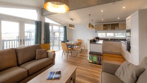 Una sala de estar moderna con un sofá, una mesa de centro y un comedor. Se ve una cocina al fondo con armarios de madera y vistas al exterior.