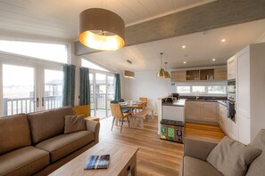 Ein modernes Wohnzimmer mit Sofa, Couchtisch und Essbereich. Im Hintergrund ist eine Küche mit Holzschränken und Blick ins Freie zu sehen.