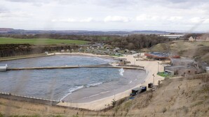 Una zona costera con una gran masa de agua, una playa de arena y varios edificios, incluyendo una estructura grande con techo plano.