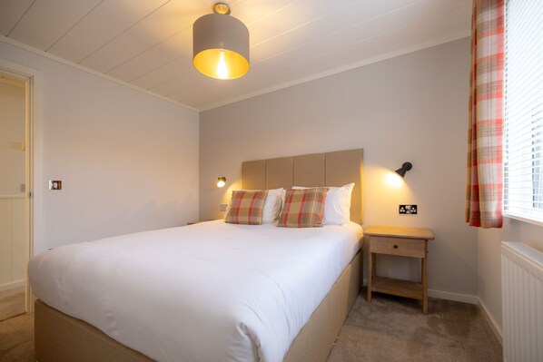 A neatly made bed with a headboard, a bedside table with a lamp, and a window with curtains.