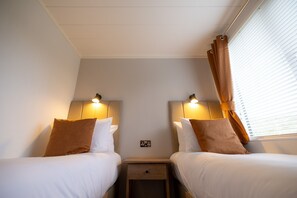 A hotel room with two beds, a nightstand, a wall socket, and a window with blinds.