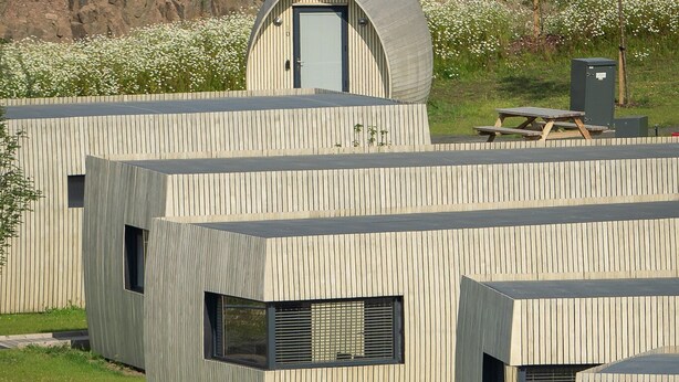 Un edificio moderno con un techo curvo distintivo, un espacio cerrado con puerta y una zona verde con gente y kayaks.