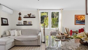 Ein Wohnzimmer mit einer Couch, einem Esstisch mit Blumenvase, einem Deckenventilator und einem Fenster mit Blick auf Grünflächen.