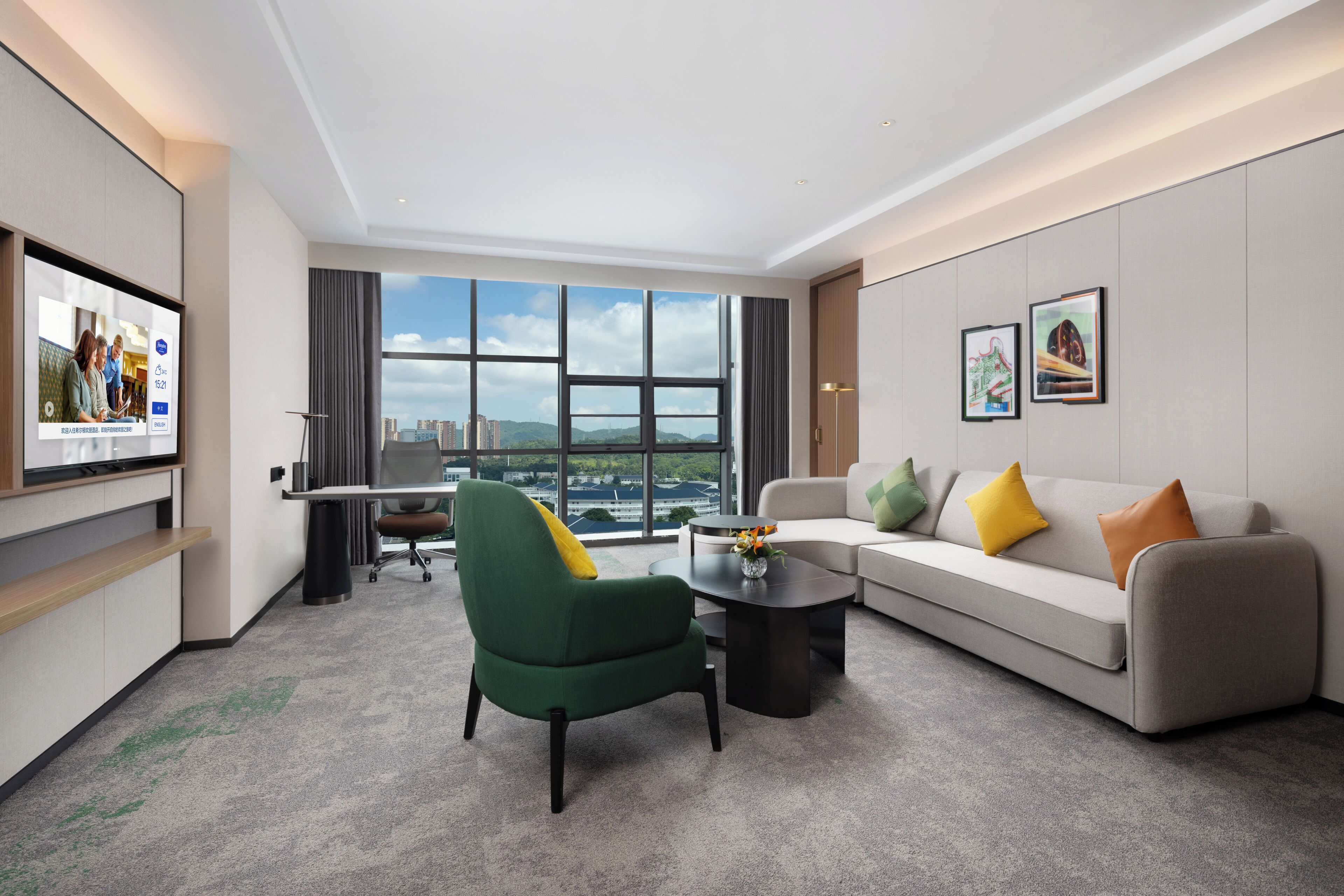 Quarto de hotel moderno com uma janela grande, um sofá, uma poltrona verde, uma mesa de centro e uma televisão de tela plana.