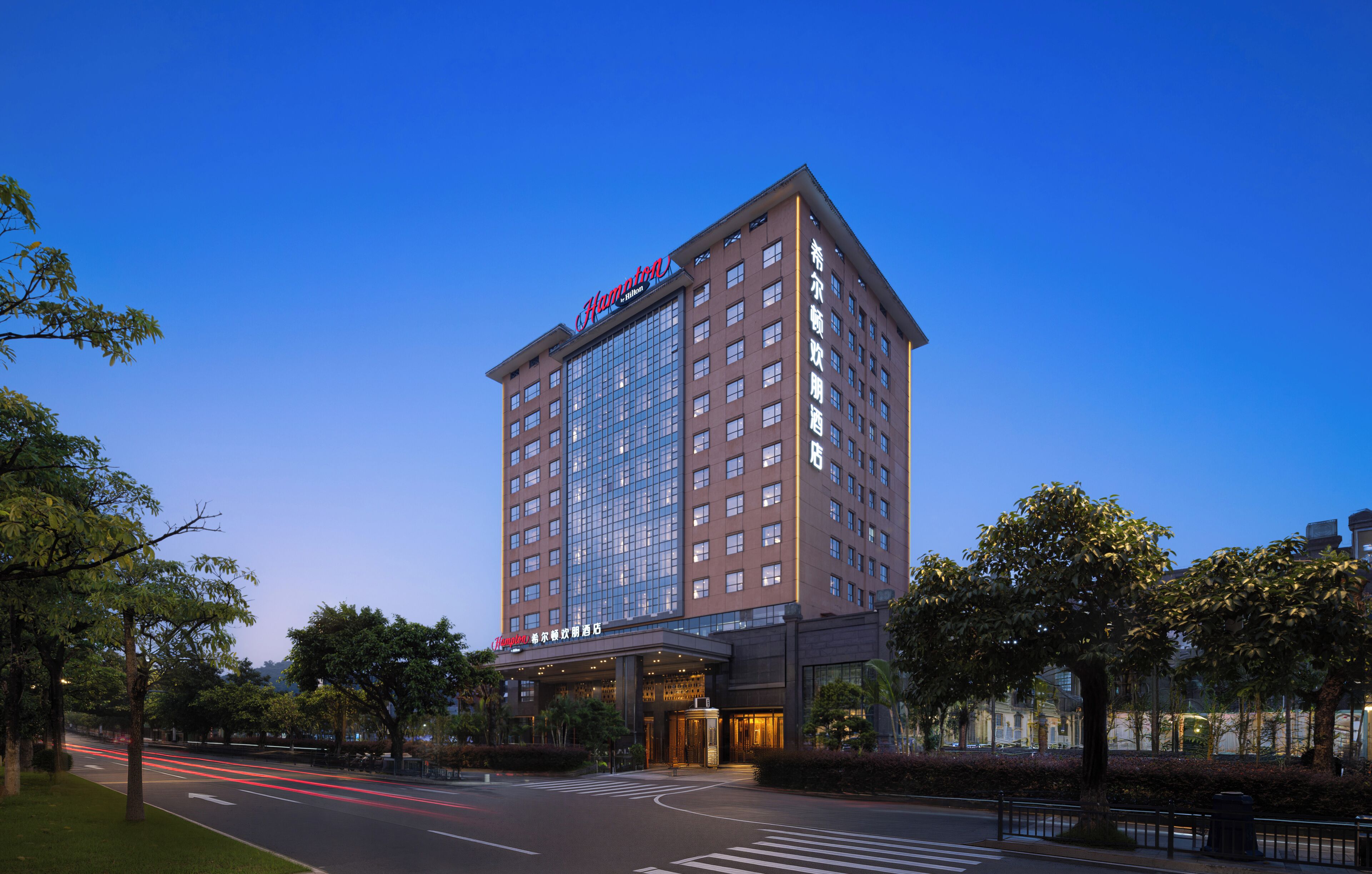 Ett högt hotell med en tydlig skylt med texten ”Hampton”, flera våningar och en modern fasad av glas.