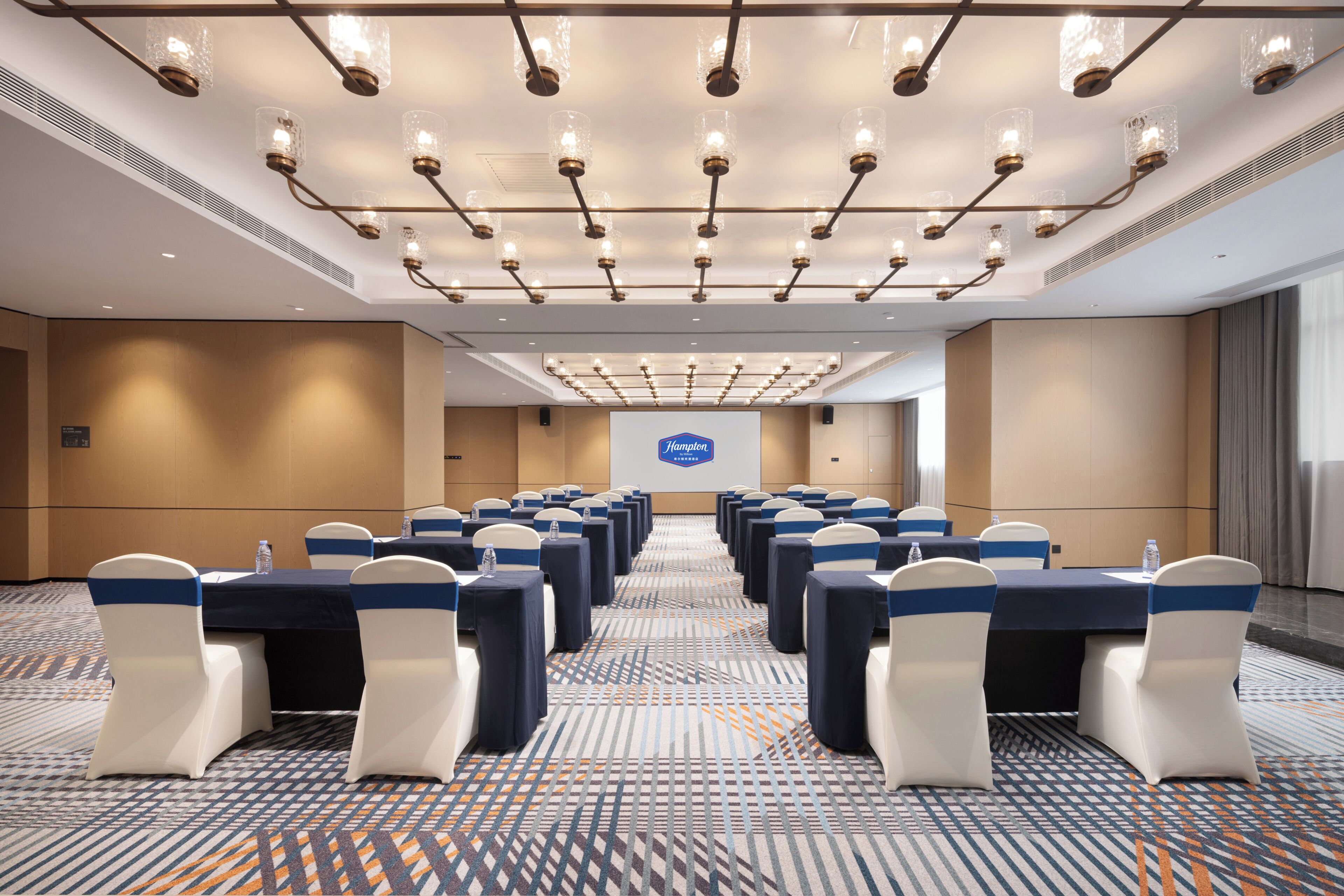 A conference room with rows of tables and chairs, a projector screen, and a patterned carpet.