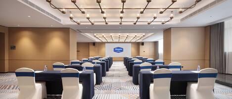 A conference room with rows of tables and chairs, a projector screen, and a patterned carpet.