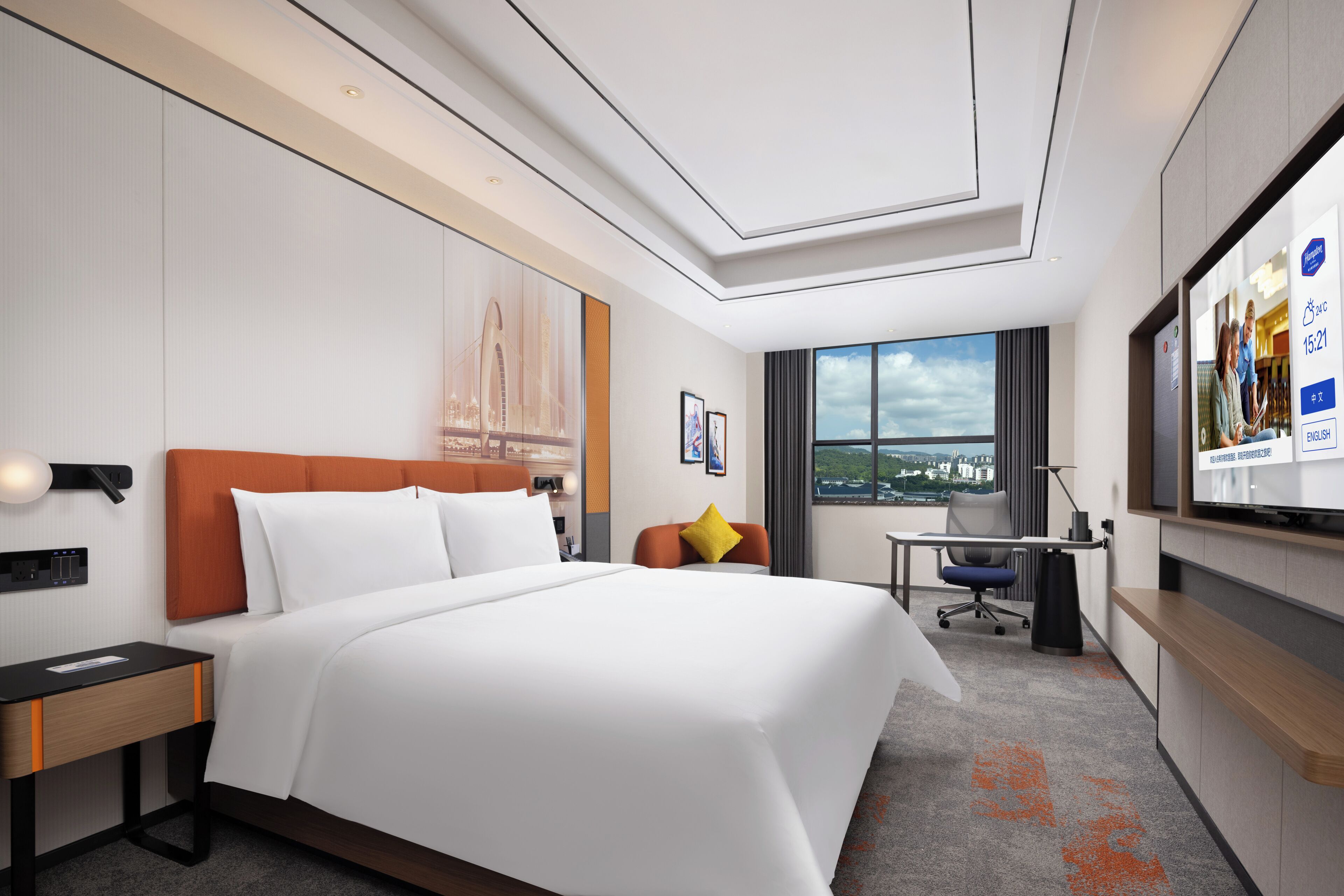 A hotel room with a large bed, a desk, a chair, a TV, and a window with a city view.
